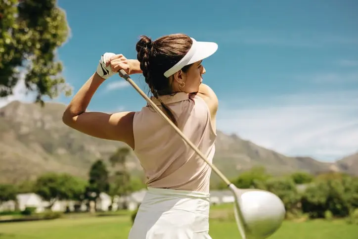 A female golfer flawlessly swinging her driver at a scenic golf resort. She is dressed in stylish sports attire, enjoying a clear, sunny day.