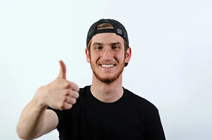 Smiling Young Adult Male in Dark T-Shirt and Baseball Hat Worn Backwards Shows His Thumbs Up
