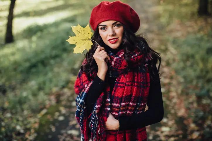 Fashionable woman wearing a red beret and plaid scarf while holding a yellow leaf in a serene autumn park, exuding warmth and modern style in a lush outdoor environment.